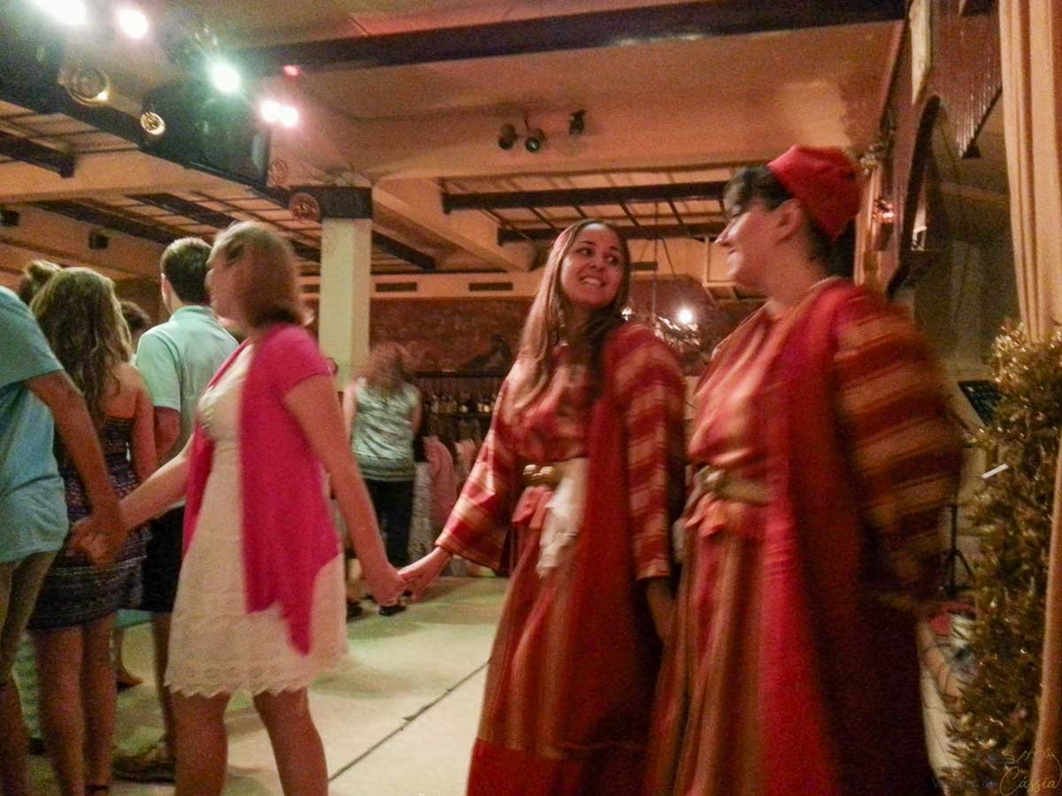 dancarinas-trajes-tipicos-taverna-atenas Dançarinas com vestidos tradicionais vermelhos sorrindo de mãos dadas com turistas na taverna.