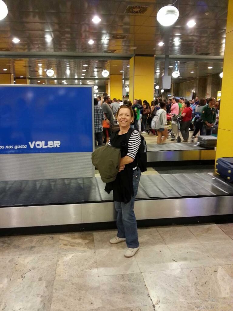 Mulher de casaco e mochila em pé ao lado da esteira de bagagens no aeroporto.