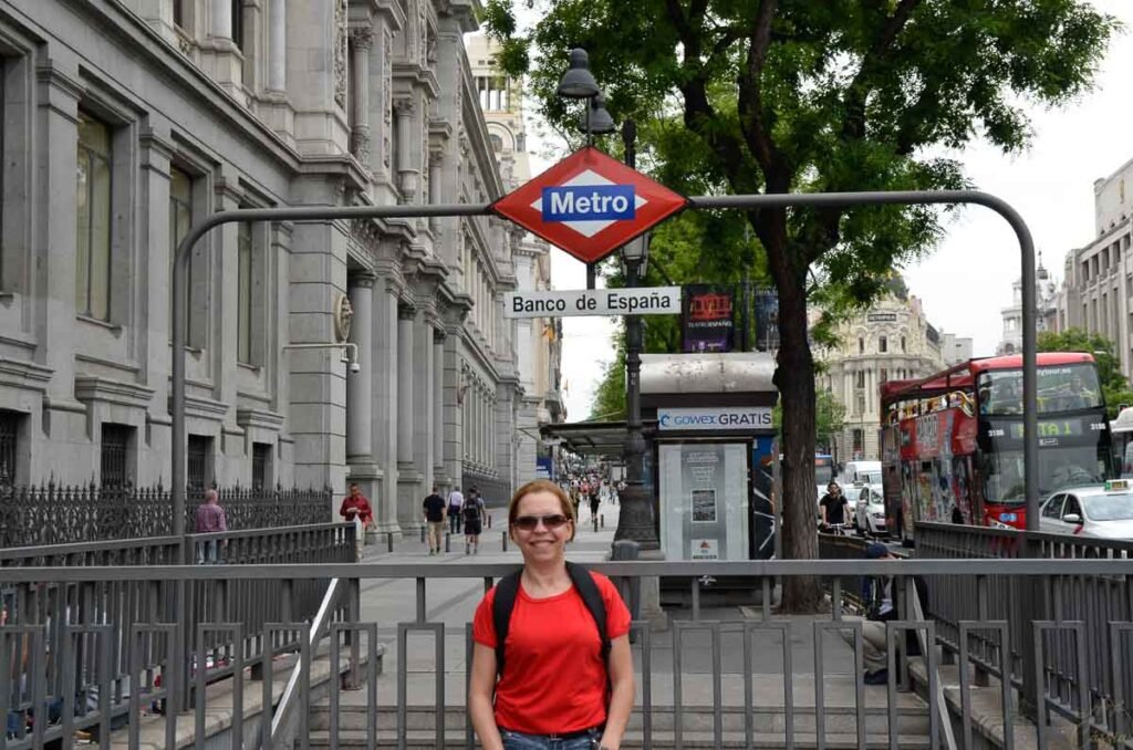 Mulher de camisa vermelha sorri em frente à entrada da estação de metrô Banco de España em Madri.