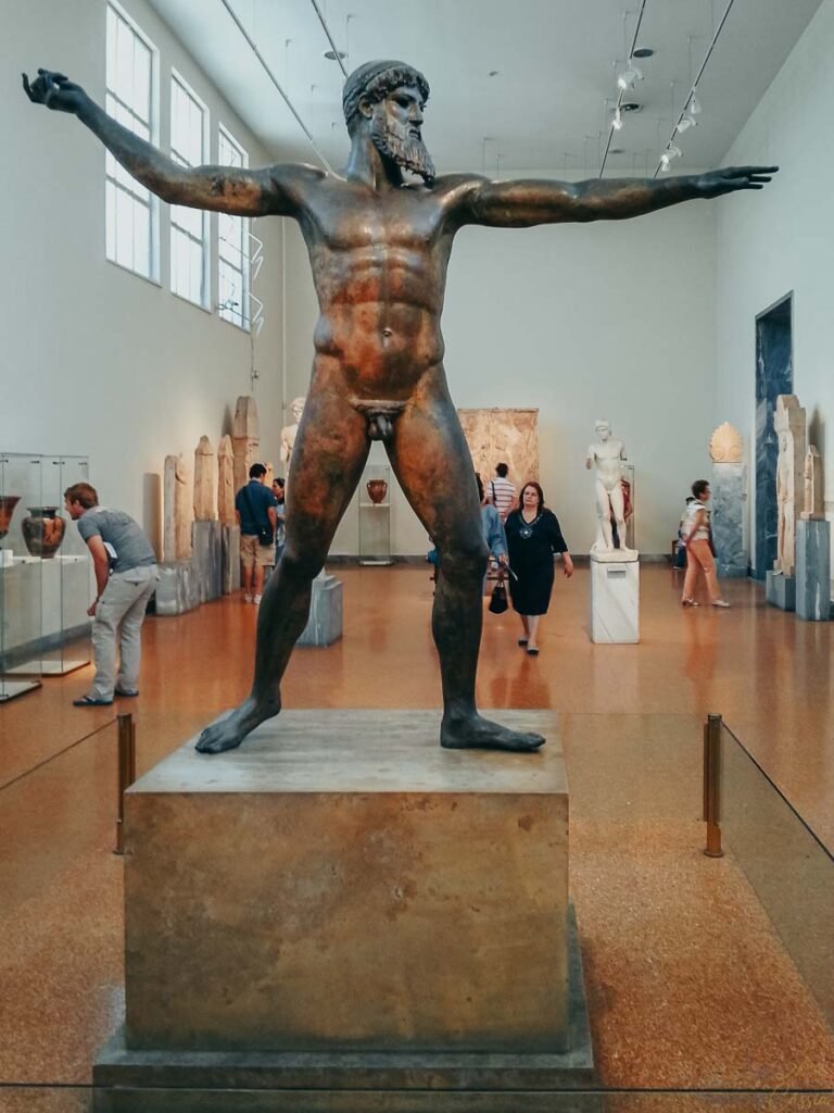 Estátua antiga de bronze de Zeus ou Poseidon em pé com braços abertos em salão de museu.