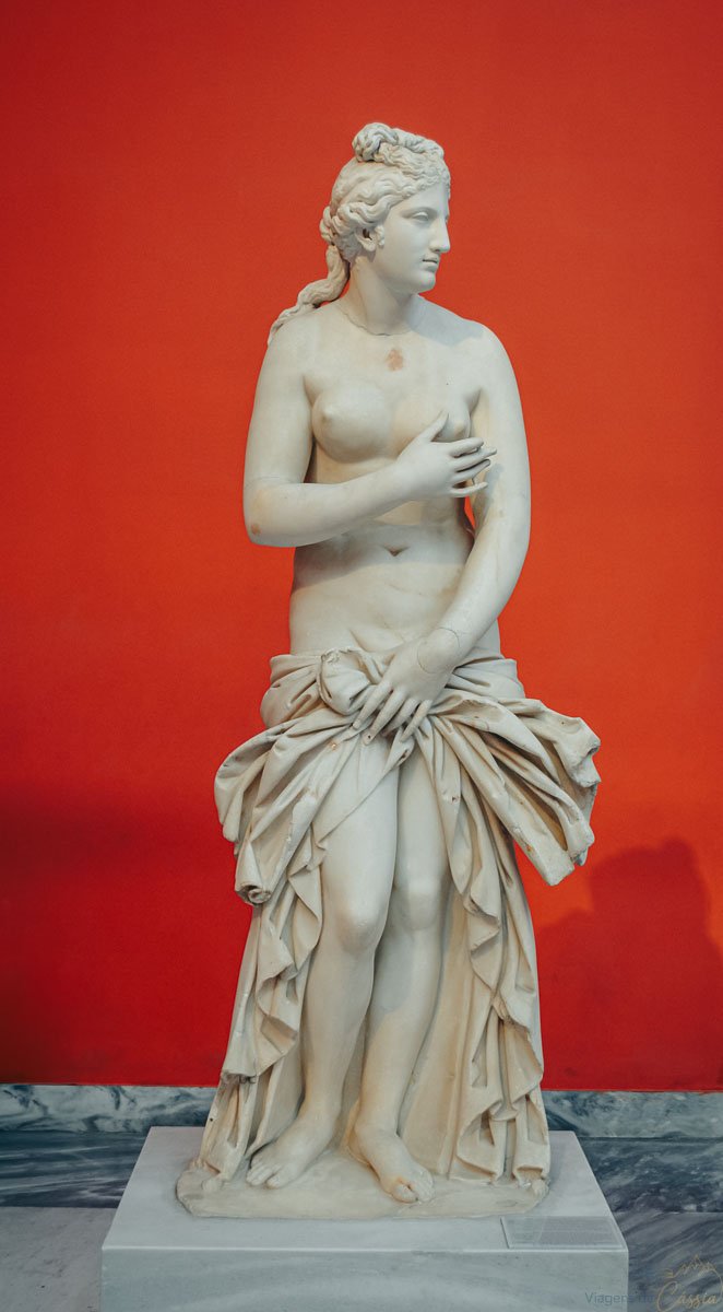 estatua-marmore-afrodite-museu-atenas Estátua de mármore branco de deusa grega em fundo vermelho vibrante no museu.