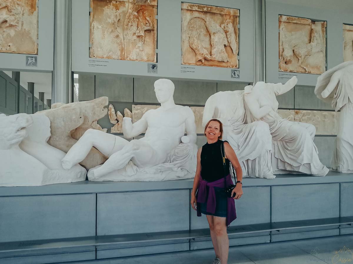 estatuas-frontao-partenon-museu-acropole Mulher sorrindo em frente às estátuas de mármore do frontão do Partenon em exposição no museu.