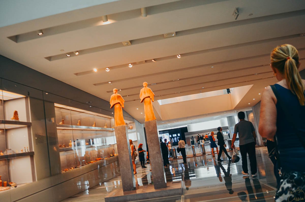 estatuas-korai-antigas-museu-acropole Esculturas arcaicas sobre pedestais altos em salão moderno com vitrines e visitantes no museu.