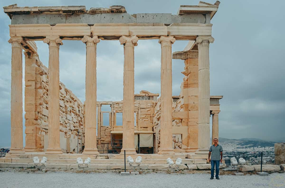 fachada-colunas-erechtheion-acropole-atenas Homem em pé diante da fachada com colunas clássicas do templo Erecteion na Acrópole de Atenas.