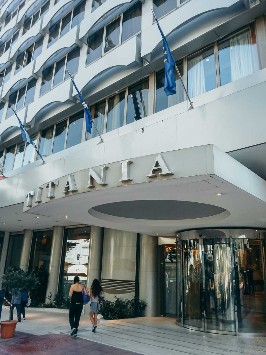fachada-entrada-hotel-titania-atenas Fachada moderna e porta giratória de entrada do Hotel Titania em Atenas com bandeiras.