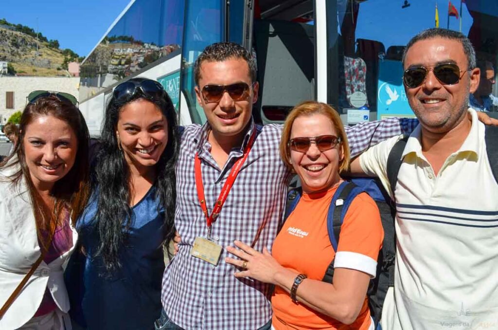 Grupo de cinco turistas sorrindo ao lado de um ônibus de viagem em um dia ensolarado.
