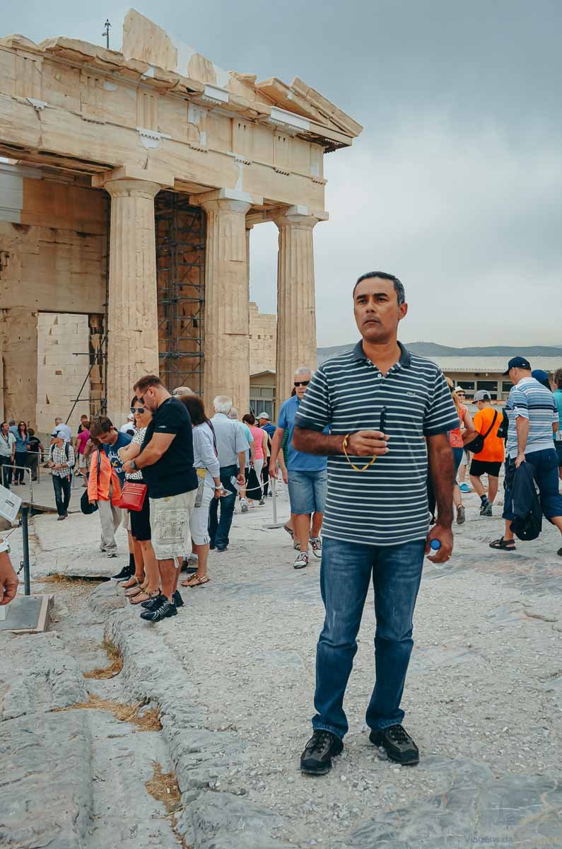 homem-ruinas-propileus-acropole-atenas Homem de camisa listrada em pé em frente às colunas monumentais dos Propileus na Acrópole de Atenas.
