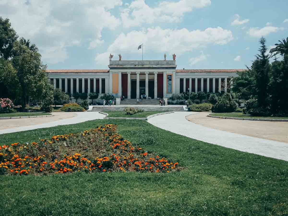 jardim-museu-arqueologico-nacional-atenas Jardins floridos com gramado verde em primeiro plano e a fachada do Museu Arqueológico ao fundo.