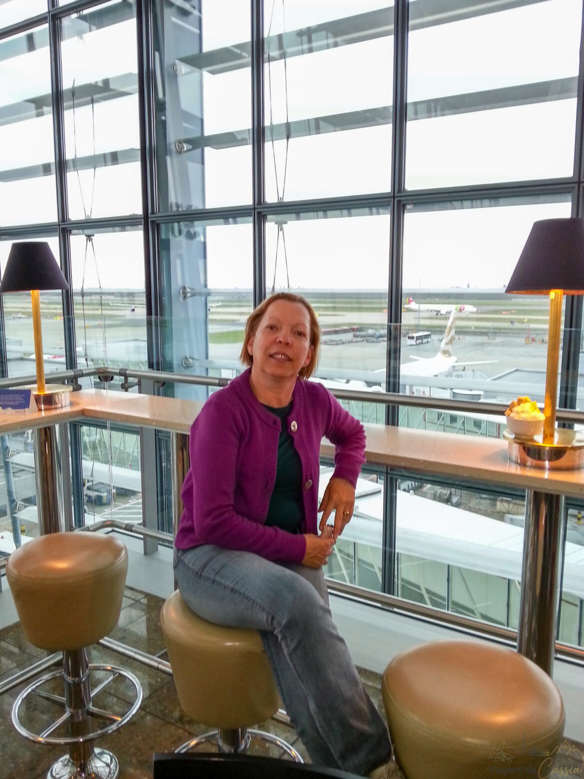 Mulher de casaco roxo sentada em banco alto perto de janelão com vista para pista de aeroporto.