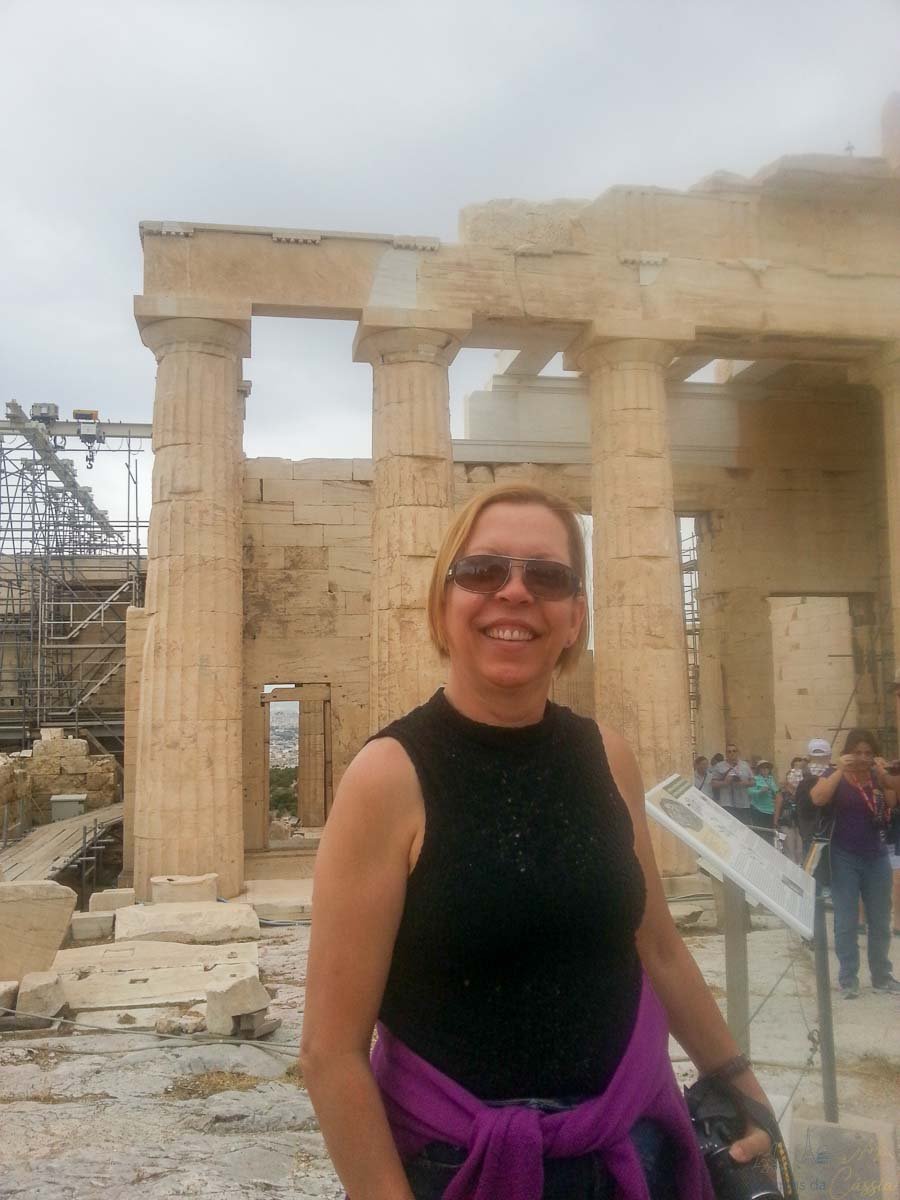 mulher-sorrindo-propileus-acropole-grecia Mulher de óculos escuros sorrindo em frente aos blocos de pedra e colunas dos Propileus em Atenas.