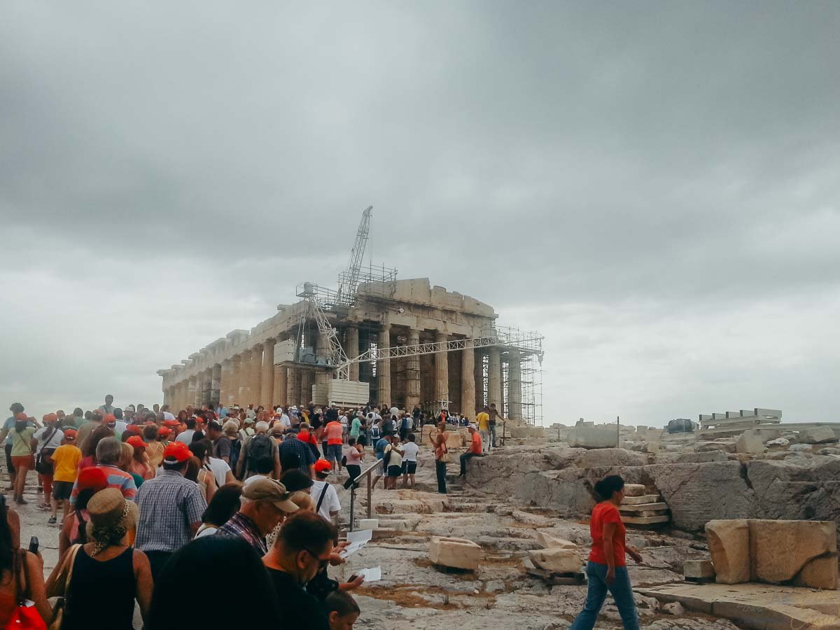 multidao-subida-partenon-acropole-atenas Multidão de visitantes caminhando em direção ao Partenon com guindastes de restauração em Atenas.