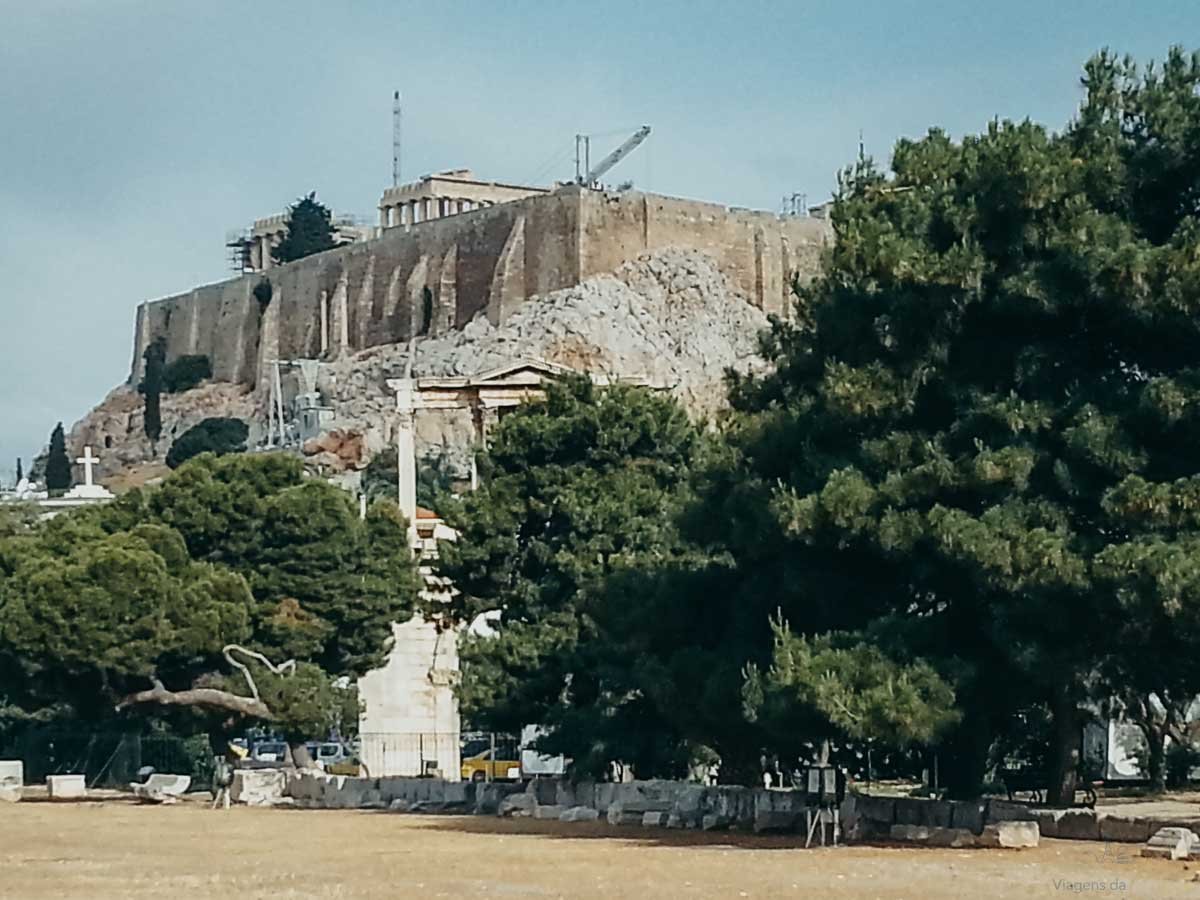 muralhas-acropole-partenon-atenas-grecia Vista inferior das muralhas de pedra da Acrópole e topo do Partenon entre árvores verdes.