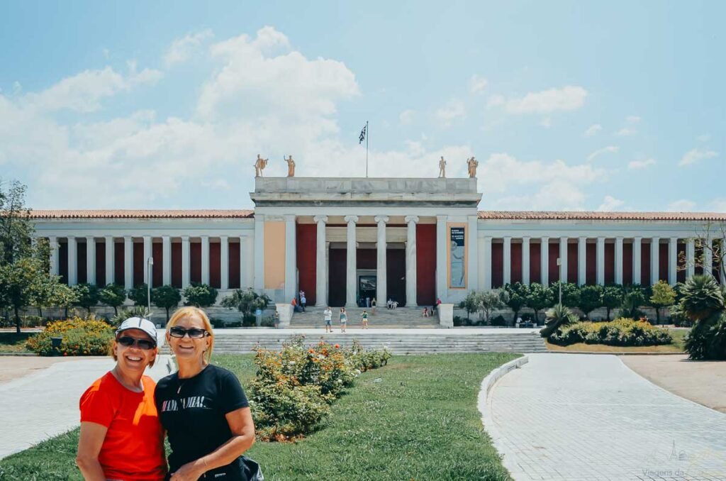 Duas mulheres sorrindo em frente à fachada neoclássica do Museu Arqueológico Nacional de Atenas.