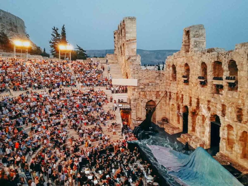Visão elevada do palco com orquestra e público nas arquibancadas do teatro de pedra.