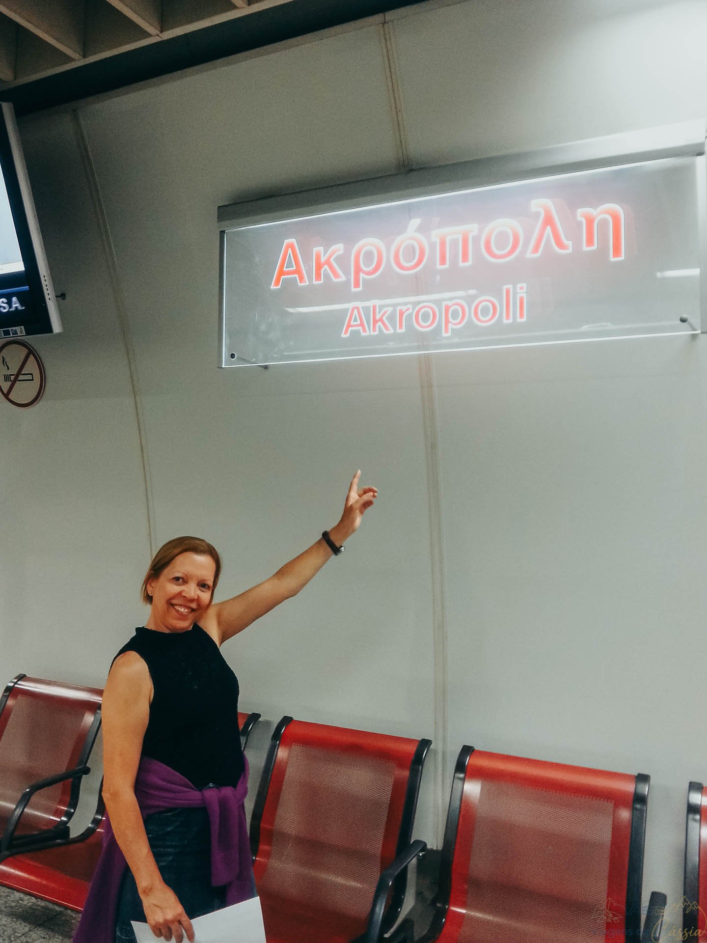 placa-estacao-metro-acropole-atenas Mulher sorrindo e apontando para a placa luminosa com o nome da estação de metrô Akropoli em Atenas.