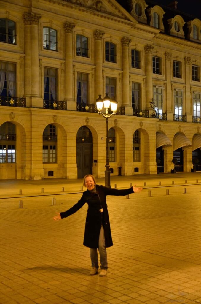 Mulher com casaco preto de braços abertos na calçada iluminada da Place Vendôme em Paris.