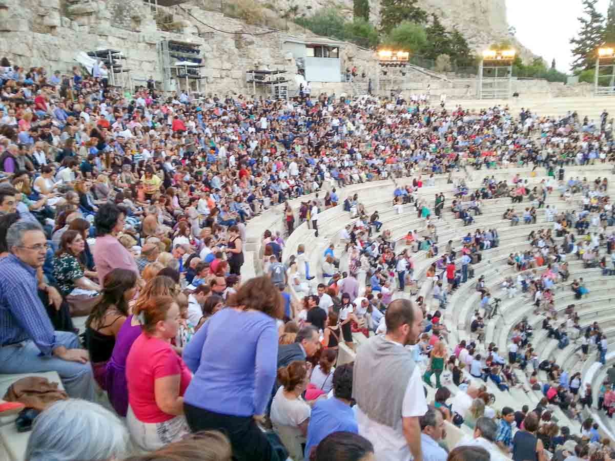 plateia-espetaculo-odeao-herodes-atico-atenas Público acomodado nas arquibancadas de pedra aguardando início de apresentação em teatro grego.