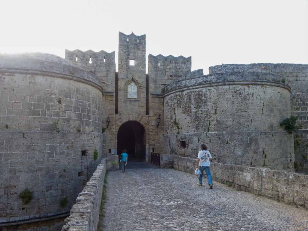 Entrada fortificada com torres circulares de pedra e ponte de acesso com pessoas caminhando.