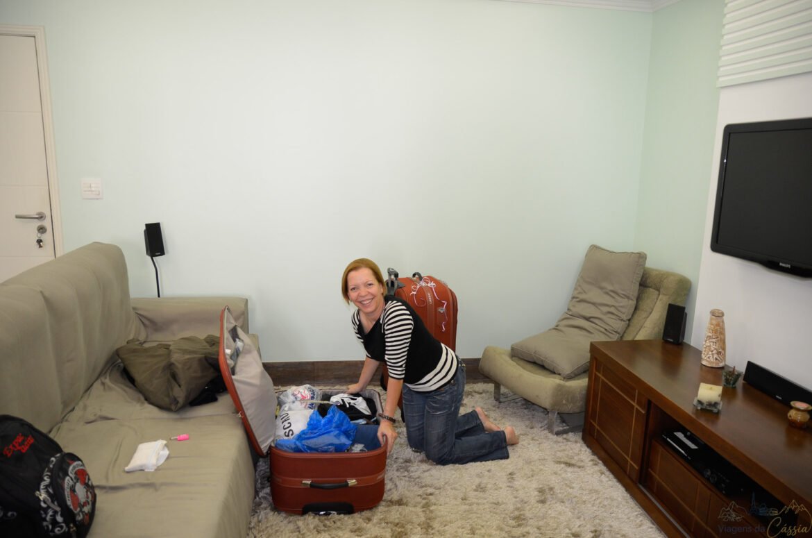 Mulher ajoelhada no tapete arrumando mala vermelha aberta em uma sala.