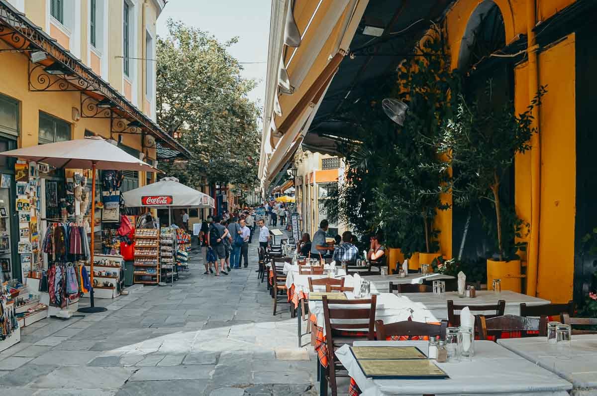 restaurantes-mesas-rua-plaka-atenas Rua de pedestres em Plaka com mesas de restaurantes ao ar livre sob ombrelones.