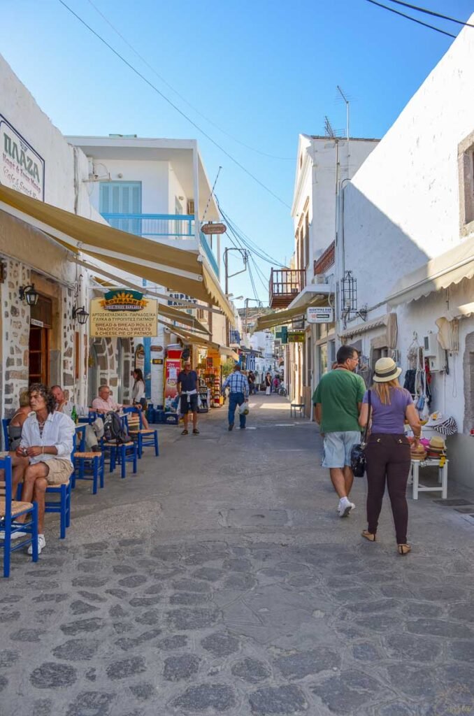 Rua de paralelepípedos com lojas tradicionais, toldos e turistas caminhando em vila grega.
