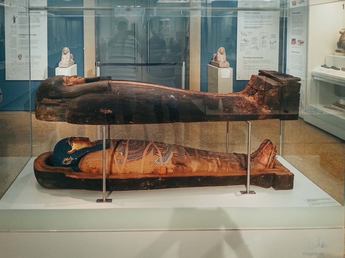 sarcofago-mumua-egipcia-museu-atenas Sarcófago de madeira entalhada e múmia egípcia preservada expostos em vitrine de vidro.