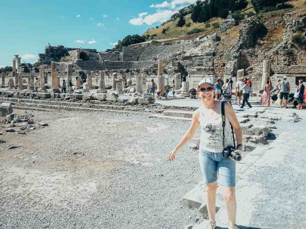Mulher sorrindo na praça antiga de pedra com colunas e arquibancadas do Odeion ao fundo.