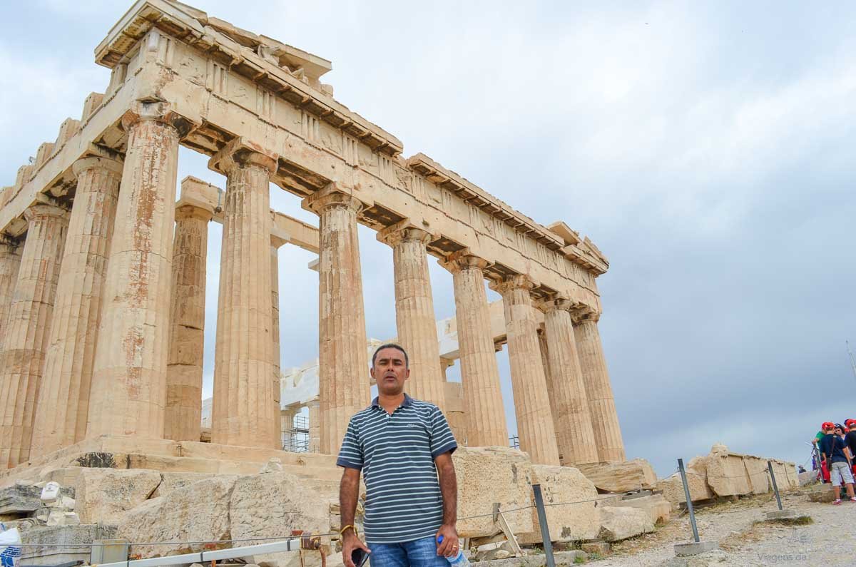 turista-base-partenon-acropole-grecia Homem posando em ângulo inferior diante da monumental fachada de colunas dóricas do Partenon.