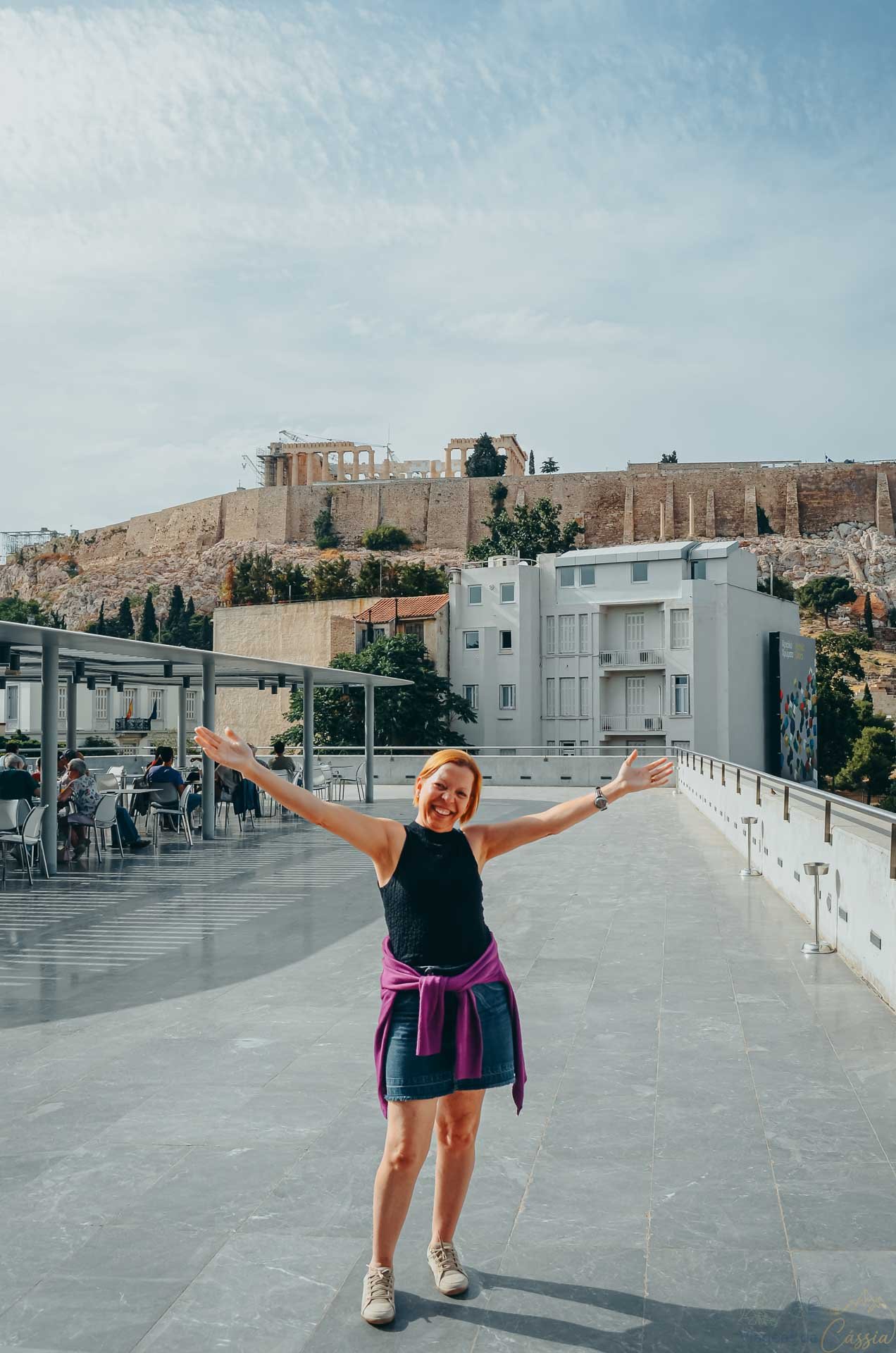 turista-bracos-abertos-vista-acropole-museu Mulher de braços abertos no terraço panorâmico com a colina da Acrópole ao fundo.