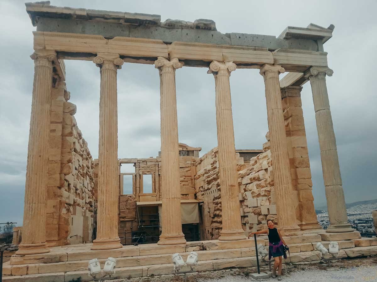 turista-colunas-erectonion-acropole-atenas Mulher com casaco roxo na cintura apoiada no guarda-corpo diante das colunas do Erecteion.