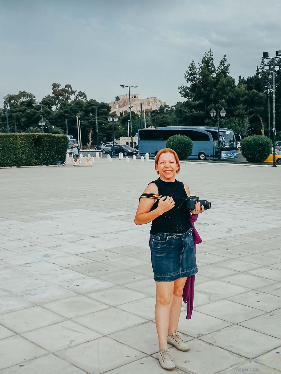 turista-com-camera-vista-acropole-atenas Mulher sorrindo segurando câmera fotográfica com a colina da Acrópole e o Partenon ao fundo.