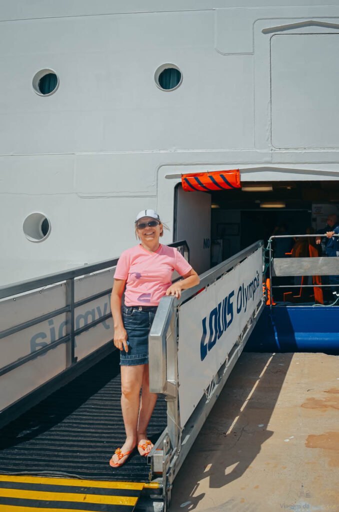 Mulher de camiseta rosa sorri na passarela de embarque de um grande navio de cruzeiro branco.