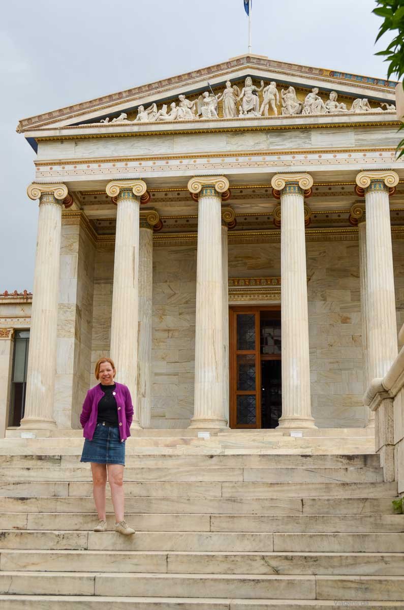 turista-escada-entrada-academia-de-atenas Mulher de casaco roxo em pé na escadaria de mármore da Academia de Atenas.