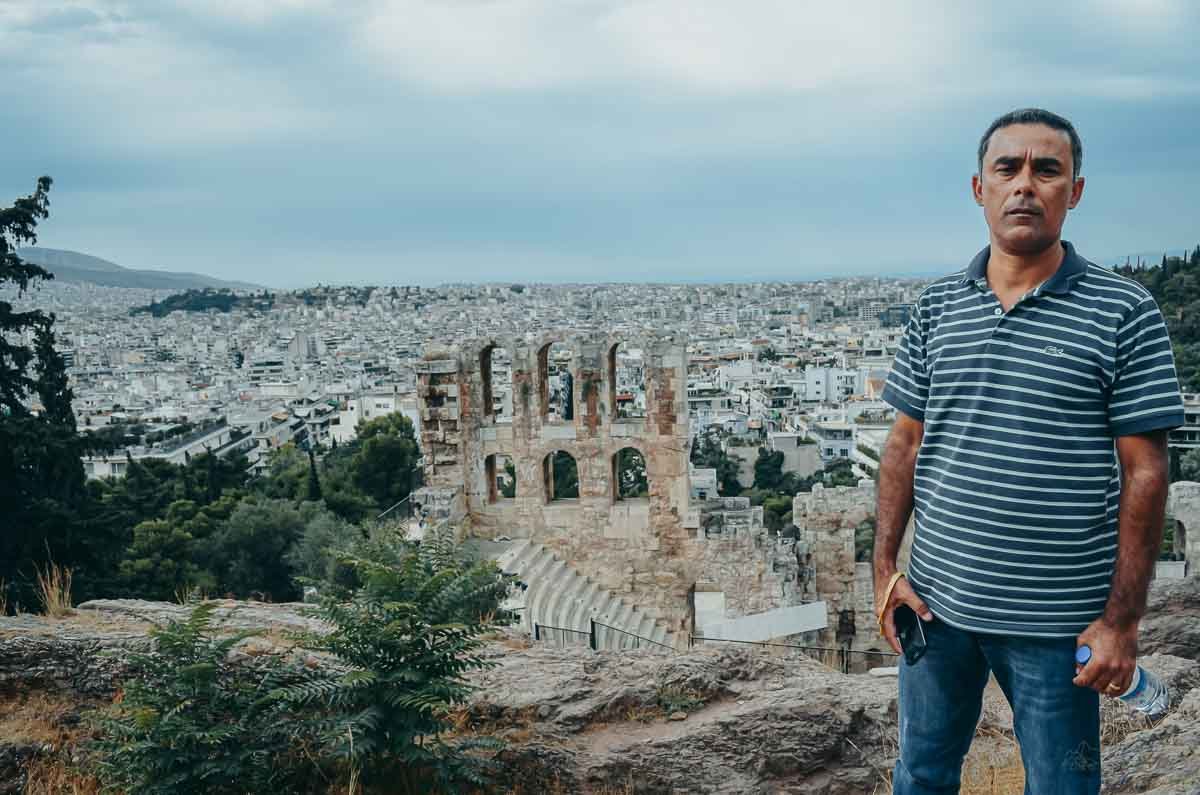 turista-frente-odeao-herodes-atico-atenas Homem de camisa polo listrada posando no mirante com arcos do Odeão e cidade ao fundo.