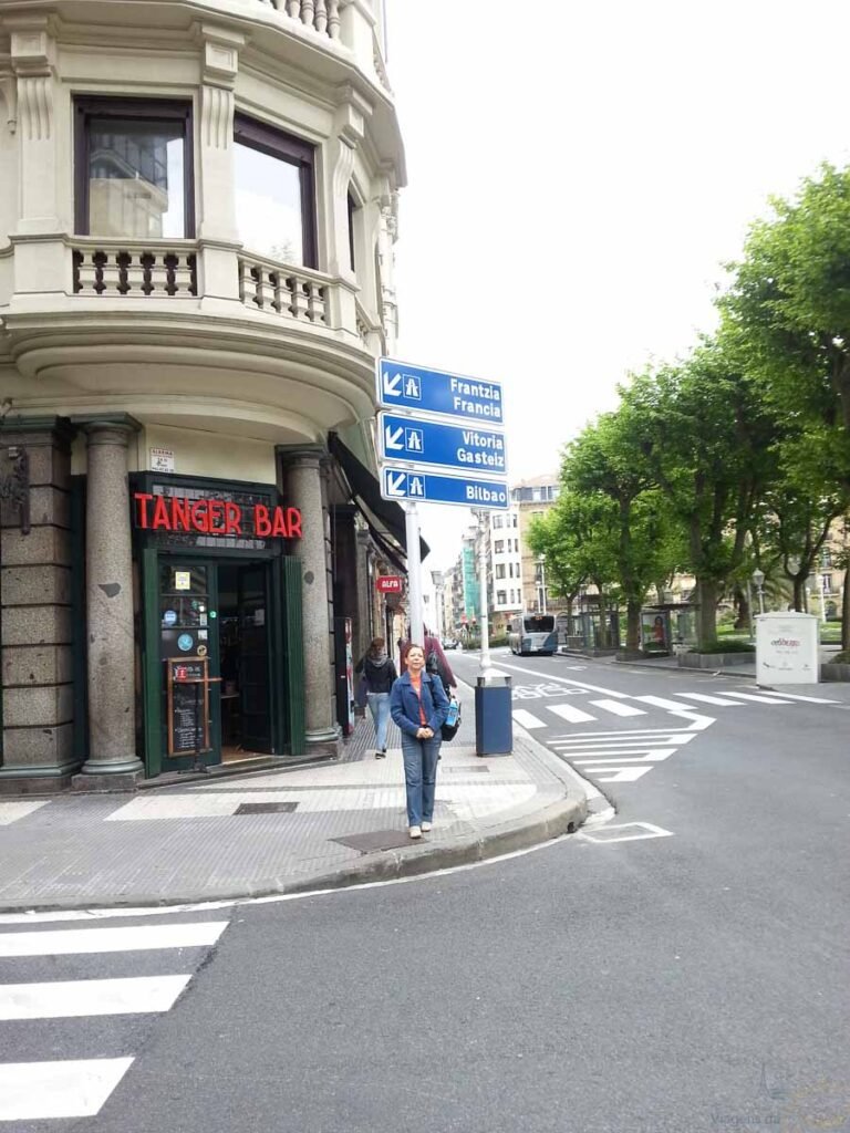 Mulher de casaco azul em pé na calçada em frente a um bar e placas indicativas de trânsito.