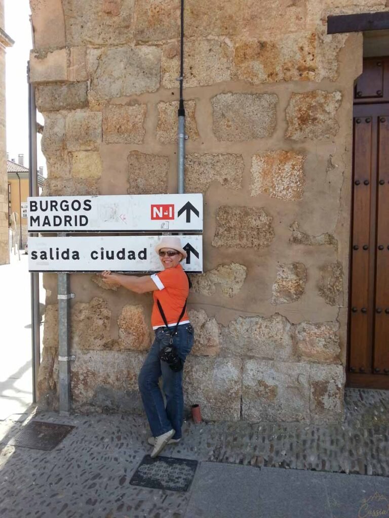 Mulher de chapéu segurando placa de trânsito com indicações para Burgos e Madrid.