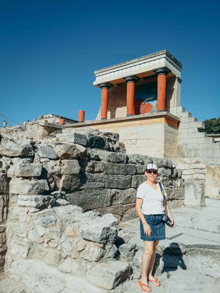 Mulher em pé sobre pedras antigas com o Bastião Norte de Cnossos ao fundo sob céu azul.