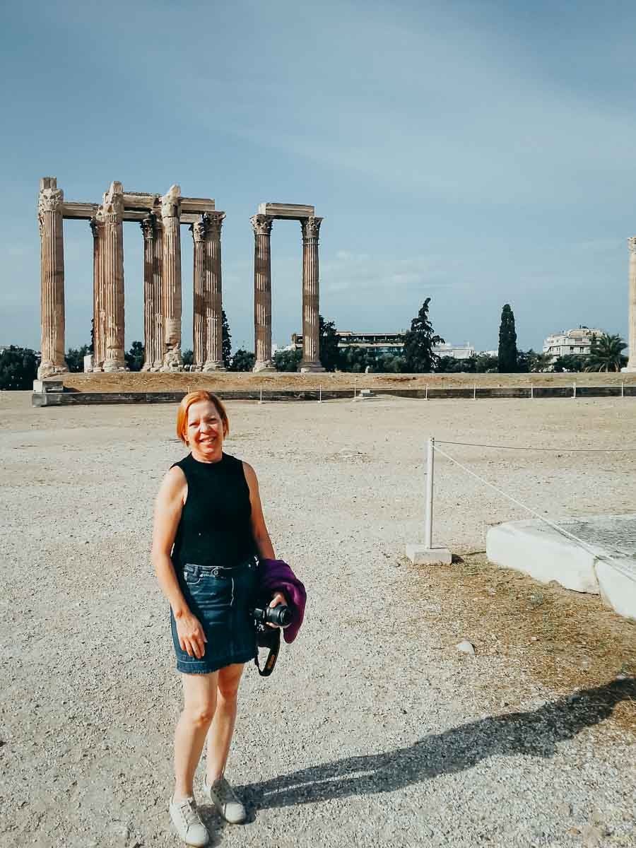 turista-ruinas-colunas-templo-zeus-olimpico Mulher com casaco roxo amarrado na cintura sorrindo em frente às colunas do Templo de Zeus.