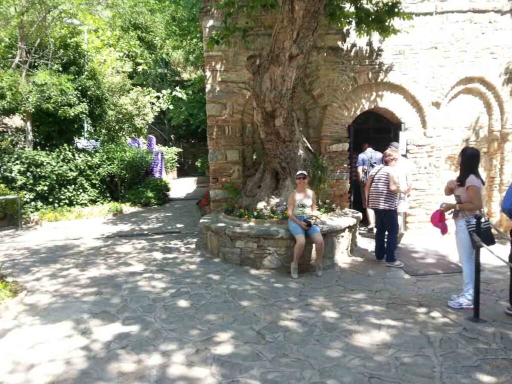 Mulher sentada sob grande árvore histórica em frente à capela da Casa da Virgem Maria.