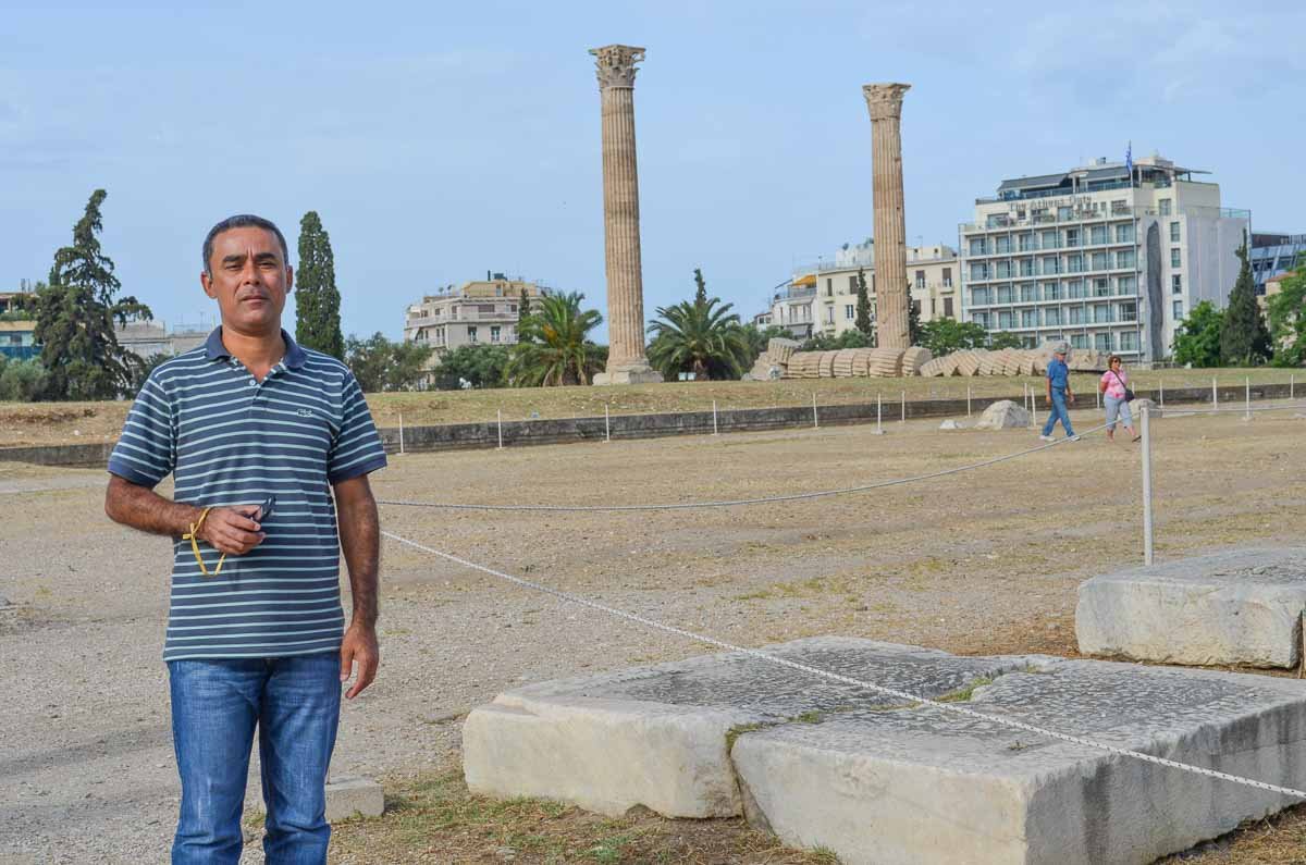 turista-sitio-arqueologico-templo-zeus-atenas Homem em pé diante das colunas históricas e ruínas do Templo de Zeus Olímpico em Atenas.