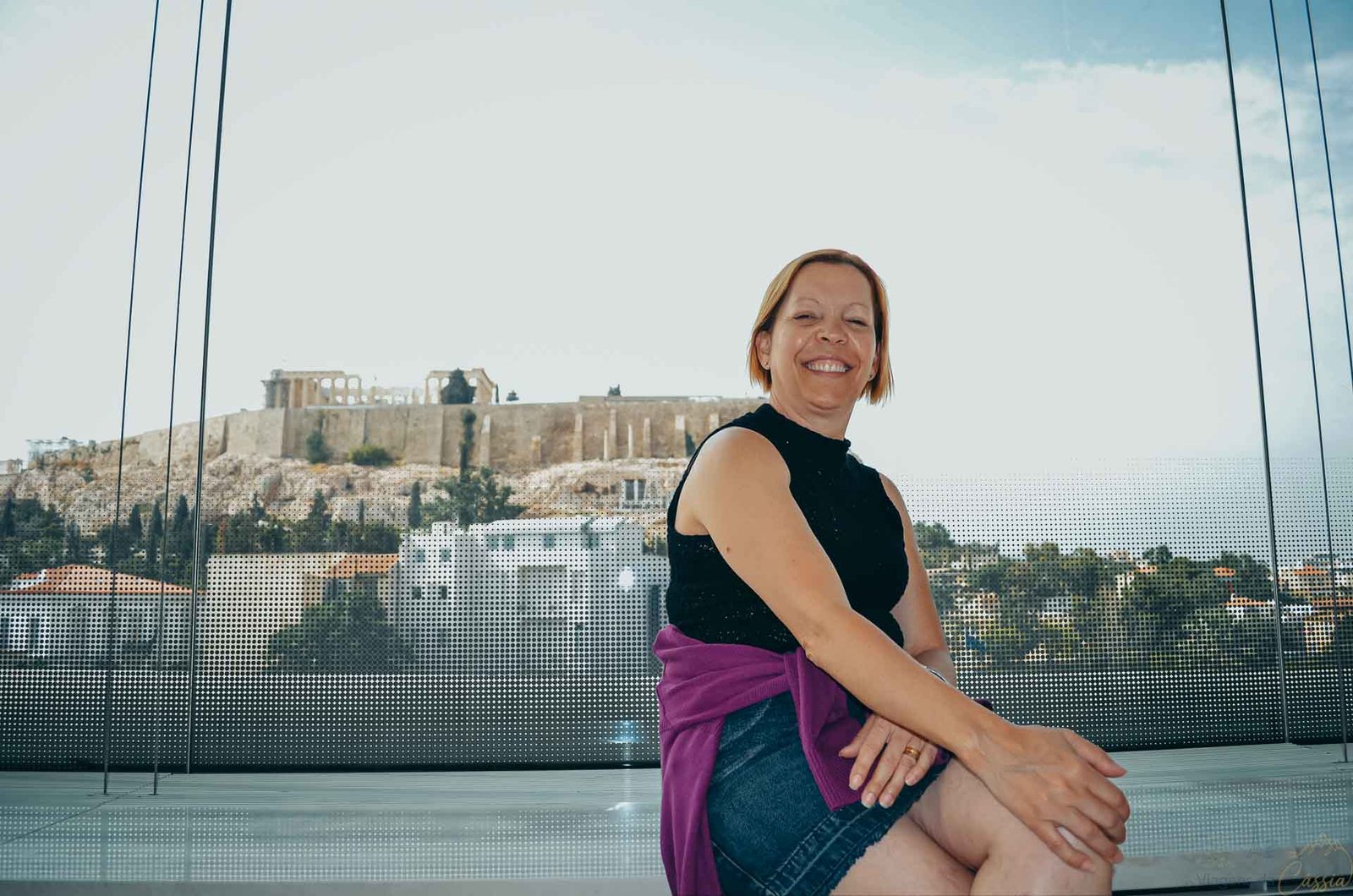 turista-terraco-museu-acropole-atenas Mulher sentada no terraço de vidro do Museu da Acrópole com vista direta para a colina histórica.