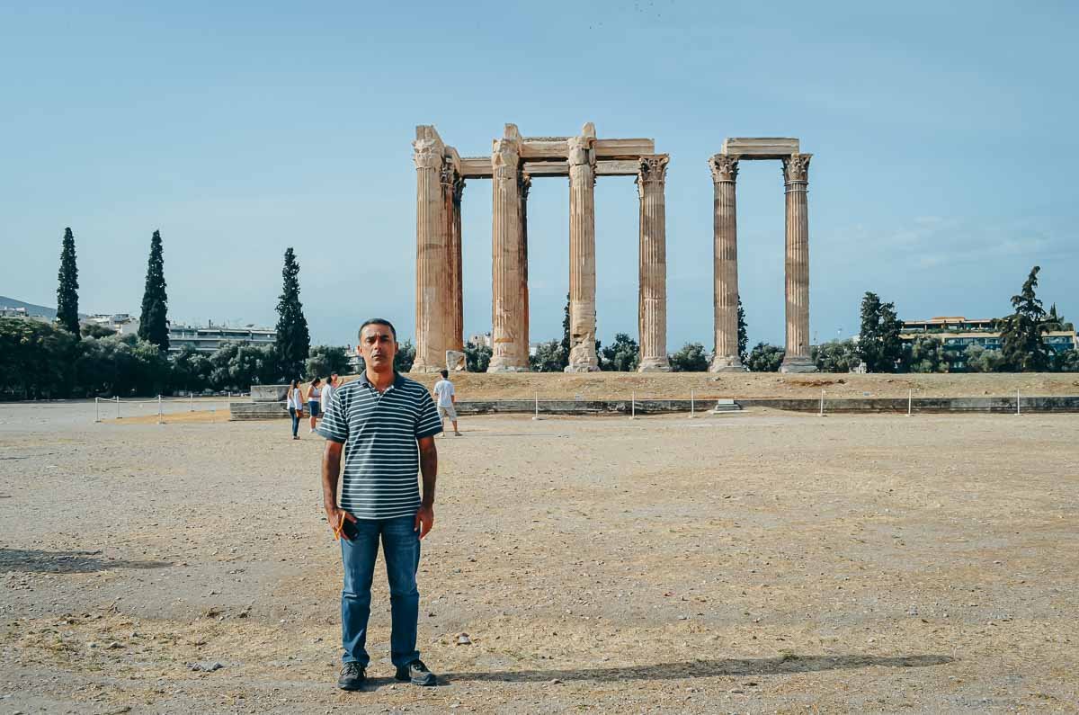visitante-terreno-templo-zeus-olimpico-grecia Homem posando no amplo pátio de terra em frente à estrutura do Templo de Zeus Olímpico.